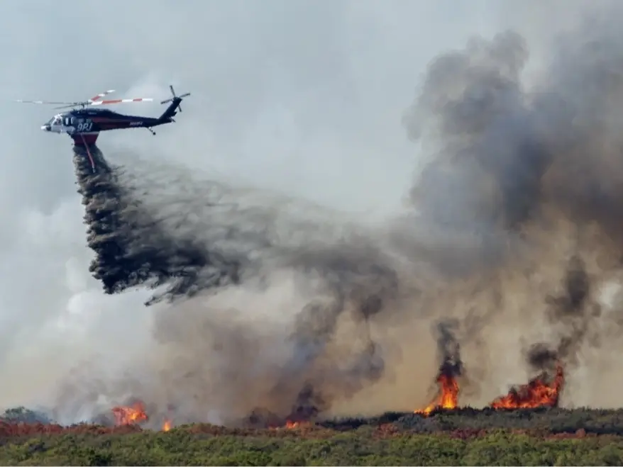 A firefighting helicopter makes a low, slow water drop directly onto flames and heavy smoke rising from a ridge of burning brush.